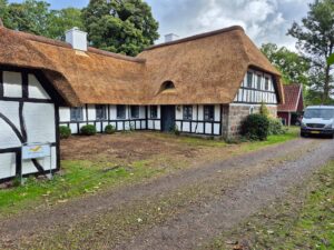 Facaden af et stort, hvidt bindingsværkshus med færdiglagt stråtag og en lille kvist.
