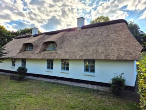 Facade af det hvide hus med stråtag og kviste, set fra haven i blødt aftenlys.