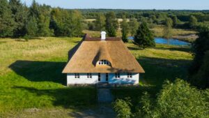 Luftfoto af huset med stråtag set forfra, beliggende i et åbent landskab med en å i baggrunden.